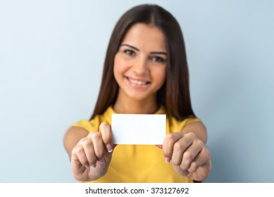 Photo Of Beautiful Young Business Woman Standing Near Gray Background. Woman With Yellow Shirt Looking At Camera, Smiling And Showing Visit Card. Focus On Visit Card