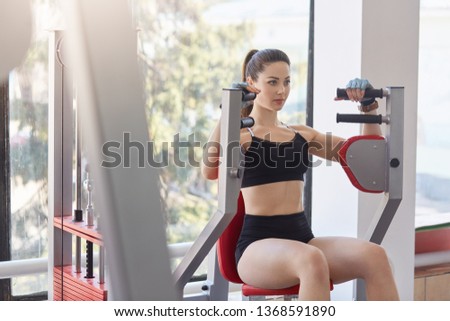 Photo of beautiful young girl performing weight lifting to improve her upper body on machine in gym. Concentrated girl has workout, tones muscles, develops strength, gains muscle mass. Sport concept.