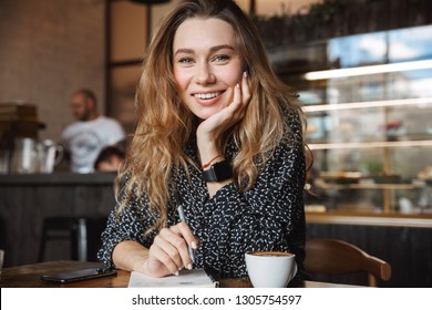 Foto von schönen jungen hübschen Frau sitzend in Café drinnen Noten in Notebook trinken Kaffee. Schaukamera.