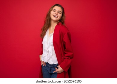 Photo of beautiful young woman with long dark hair standing wearing white t-shirt and red cardigan isolated over red background looking at camera smiling and poising hands in jeans pockets