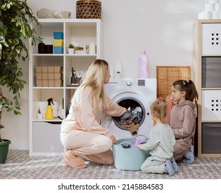 Photo From Behind Hangers With Clothes, View Of Laundry Room, Bathroom Mother With Two Daughters Kneels Next To Washing Machine By Bowl With Clothes Women Throw Dirty Things Into Drum Spinning Drying.