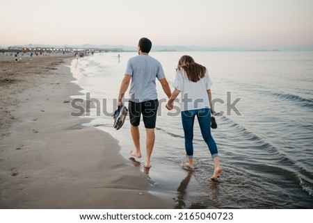 A photo from behind of a man and a woman who are fooling around the sandy beach barefoot at twilight in 
Spain. A couple of tourists on a date near the Balearic sea in Valencia.