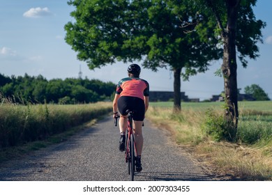 Photo From Behind Of A Woman Cyclist, Riding A Road Bike On A Road In The Middle Of Nature. Wearing A Pink And Green T-shirt