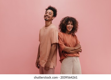photo below belt of cheerful people of exotic appearance on isolated nacre pink wall. brunette with her arms crossed dressed in carrot-colored T-shirt tucked in jeans. guy in peach t-shirt. - Shutterstock ID 2039564261