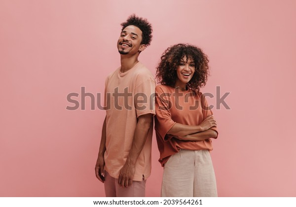 photo below belt of cheerful people of exotic\
appearance on isolated nacre pink wall. brunette with her arms\
crossed dressed in carrot-colored T-shirt tucked in jeans. guy in\
peach t-shirt.