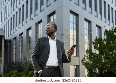 Photo from below. Portrait of an African-American businessman standing in a suit outside an office center and holding a phone. He keeps his hand in his pocket, confidently looks to the side, smiles.