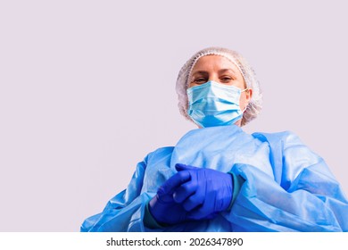 Photo From Below Of A Senior Female Doctor In A Medical Gown, Face Mask And Protective Blue Gloves Looking Down Into The Camera With Smiling Eyes.