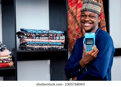 Photo of a black african man standing beside a fabric closets while holding and paying with a pos machine in a textile store