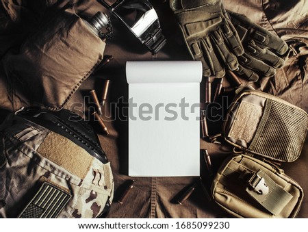 Photo of a blank notebook laying on military table with helmet, glasses, bullet shells, gloves and flask upper view.
