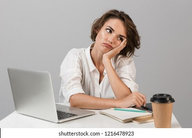 Photo Of A Bored Tired Young Woman Sitting At The Table Isolated Over Grey Background Near Laptop Computer.