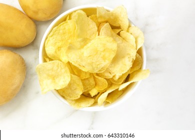 A photo of a bowl of potato chips with potatoes, shot from above on a white marble texture with a place for text