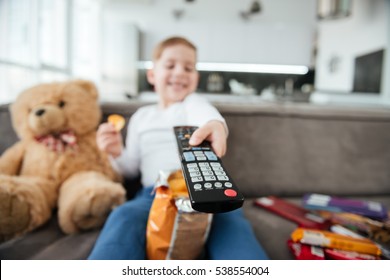 Photo of boy sitting on sofa with teddy bear at home and watching TV while eating chips. Holding remote control. Focus on remote control. - Powered by Shutterstock