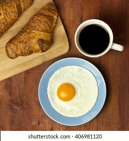 A Photo Of A Breakfast: A Perfectly Round Sunny Side Up Egg With A Cup Of Black Coffee And Croissants On A Cutting Board, Shot From Above On A Brown Wooden Texture