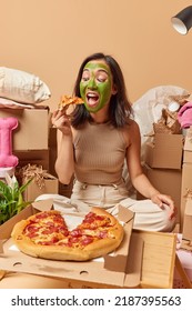 Photo Of Brunette Asian Woman Applies Green Facial Mask Eats Appetizing Pizza Sits On Floor Around Cardboard Boxes Relocates To New Place Of Living Packs Personal Belongings. Ownership Concept