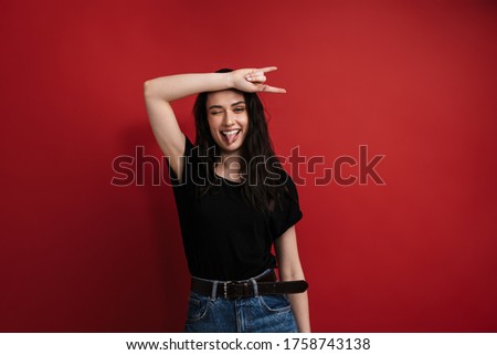 Photo of brunette funny woman in casual t-shirt winking and showing horn sign isolated over red background