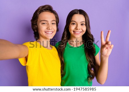 Photo of brunette happy boy and girl wear green yellow t-shirt make selfie v-sign isolated on purple color background