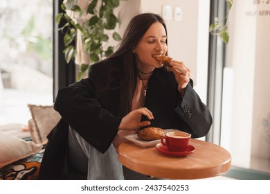 Photo of brunette woman ordering coffee and croissant. Young woman having breakfast with croissant and cup of coffee at the cafe. Girl bite croissant and look happy, enjoying of delicious meal. - Powered by Shutterstock