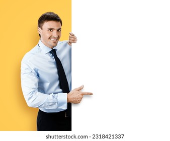 Photo of business man, employee, professional in confident shirt wear. Businessman stand behind, hang over, show point finger empty white banner signboard. Isolated against yellow background. - Powered by Shutterstock