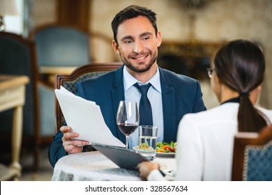 Photo Of Business Meeting In Expensive Hotel. Young Smiling Businessman Talking With Business Woman And Showing Documents While Having Dinner