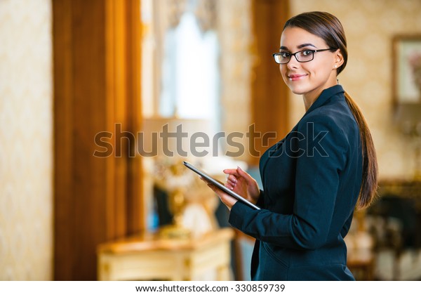 Photo of business woman in\
expensive hotel. Young business woman wearing suit and glasses,\
standing in nice hotel room, using tablet computer and looking at\
camera
