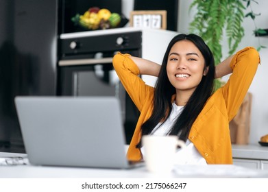 Photo Of Calm Peaceful Young Chinese Woman, Sitting At A Table In The Kitchen, Crossed Hands Behind Head, Enjoying Break Time During Distant Online Work Or Study, Looking Aside, Dreaming, Planning