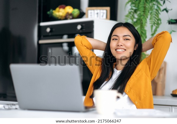 Photo of calm peaceful young chinese woman,\
sitting at a table in the kitchen, crossed hands behind head,\
enjoying break time during distant online work or study, looking\
aside, dreaming, planning