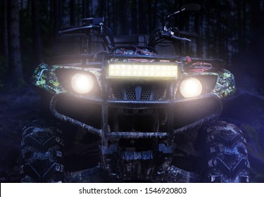 Photo of a camouflaged dirty offroad hunting atv vehicle standing at night in forest with headlights turned on, front view.