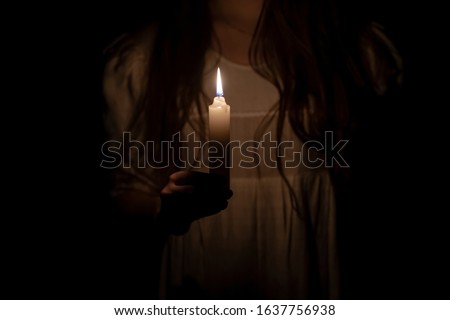 Photo of a candle at night holding by a young girl in an old white dress.  Focus on the candle. Dark background. Scary horror concept. 