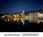 The photo captures a serene night view of a Croation harbor. Reflections of illuminated buildings and a church tower shimmer on the calm water, creating a peaceful and enchanting atmosphere.