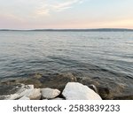 The photo captures a serne Croation coastline at sunset. Gentle waves lap against smooth stones, reflecting soft color of the sky. The distant hills add to tranquil atmosphere, inviting relaxation.