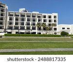 The photo captures a white hotel situated on a green clearing, surrounded by palm trees with paths for people