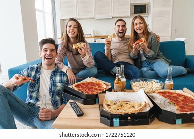 Photo of caucasian laughing friends eating pizza and drinking bear while watching tv in modern kitchen