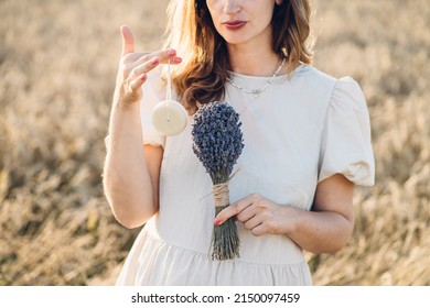 Photo Of Caucasian Pregnant Mature Woman In Dress Holding Solid Shampoo And Lavender Bouquet Of Flowers While Walking Outdoor Through Wheat Field In Summer Sunset.