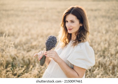 Photo Of Caucasian Pregnant Mature Woman In Dress Holding Lavender Bouquet Of Flowers While Walking Outdoor Through Wheat Field In Summer Sunset.