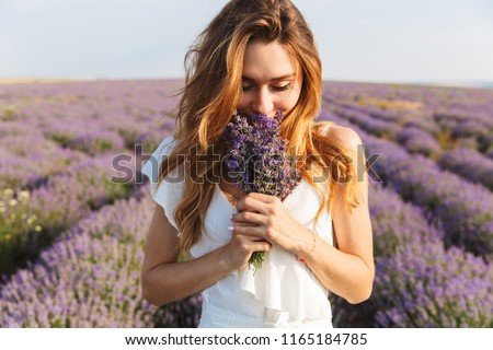 Photo of caucasian young woman in dress holding bouquet of flowers while walking outdoor through lavender field in summer