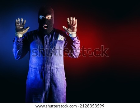 Photo of caught robber in mask, overalls, gloves and money pack standing on red and blue background.