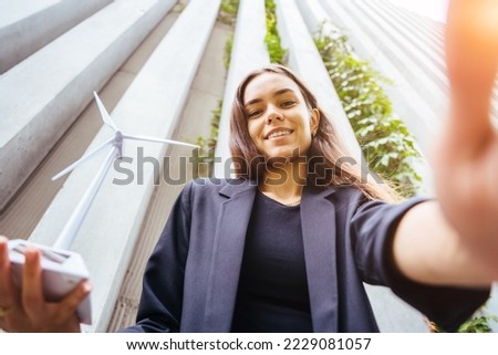 Photo of charming female student or dressed in a business suit with wind turbine model making selfie portrait outdoor. Sun glare effect.
