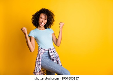 Photo of charming lucky dark skin wavy lady wear blue t-shirt rising fists empty space isolated yellow color background