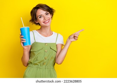 Photo of charming pretty girl wear khaki overalls drinking soda pepsi cola finger directing mockup smiling isolated on yellow color background