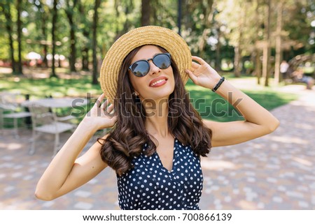 Photo of charming woman wearing summer hat and cute dress in the summer park. She is walking in the park. She holds her hat and smiles. Sunny day. Great emotions. Background green park.