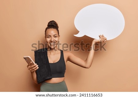 Photo of cheerful active Latin woman dressed in activewear holds speech bubble for your promotion uses smartphone checks newsfeed being in good physical shape isolated over brown background.