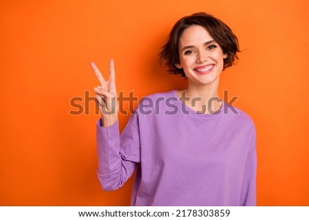 Photo of cheerful adorable girl toothy smile hand fingers demonstrate v-sign isolated on orange color background
