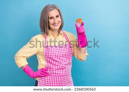 Photo of cheerful aged lady toothy smile hand hold detergent spray bottle empty space isolated on blue color background
