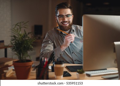 Photo Of Cheerful Bearded Web Designer Dressed In Shirt Working Late At Night And Looking At Camera While Holding Pen In Hand