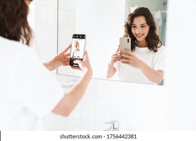 Photo of cheerful beautiful woman smiling and taking selfie on cellphone while looking at mirror at bathroom - Powered by Shutterstock