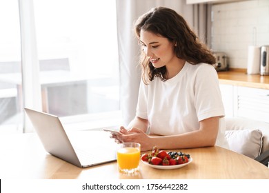 Photo of cheerful beautiful woman using laptop and mobile phone while having breakfast in modern kitchen