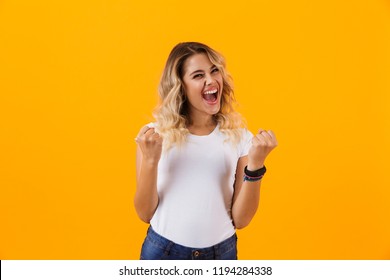 Photo of cheerful blond woman in basic clothing yelling and clenching fists isolated over yellow background