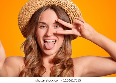 Photo of cheerful Caucasian female with funny expression, makes peace sign, shows tongue, makes selfie, wears elegant straw hat, poses against orange background. People and recreation concept - Shutterstock ID 1156617355