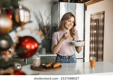 Photo of cheerful Caucasian woman with blond hair casual outfit standing in the cozy kitchen eating healthy breakfast muesli with berries with Christmas tree in the foreground front. Holidays concept - Powered by Shutterstock