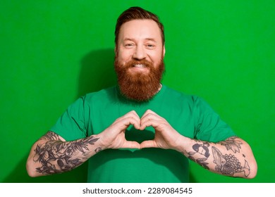 Photo of cheerful charming man wear t-shirt smiling showing arms heart isolated green color background